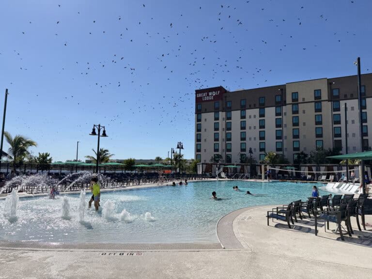 Bright outdoor hotel pool with children splashing, surrounded by lounge chairs, palm trees, and a modern building, under a clear blue sky.